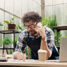 Happy Caucasian businessman talking on cellphone while standing in apron in small floral center and writing down order details. Joyful male florist calling on smartphone at work. Own business concept