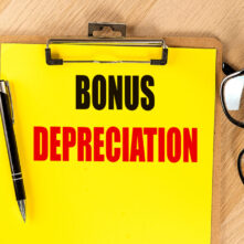 Clipboard with the text bonus depreciation, pen, and glasses on a wooden table with a yellow background